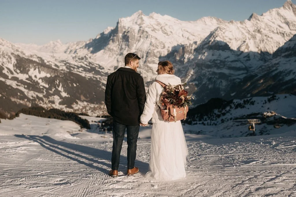 A couple in wedding attire holding hands and walking in a snowy mountain landscape with tall snow-capped peaks in the background.