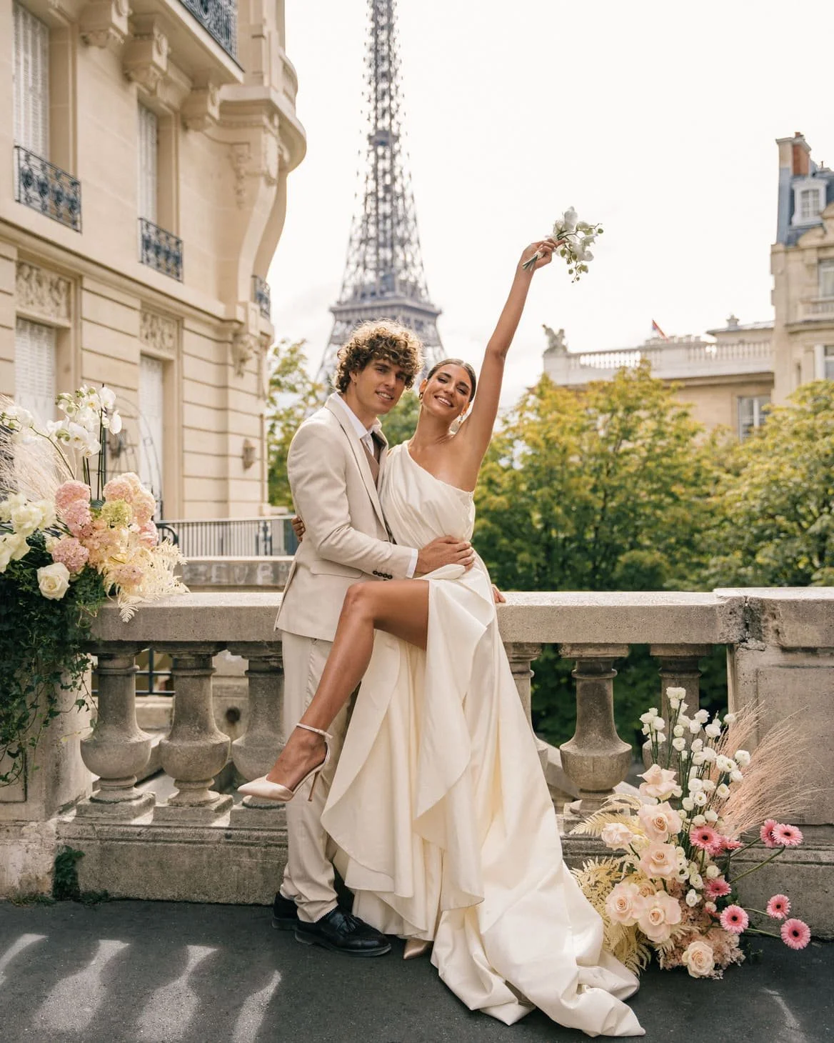 Couple in wedding attire on a Paris balcony with the Eiffel Tower in the background, surrounded by flowers.