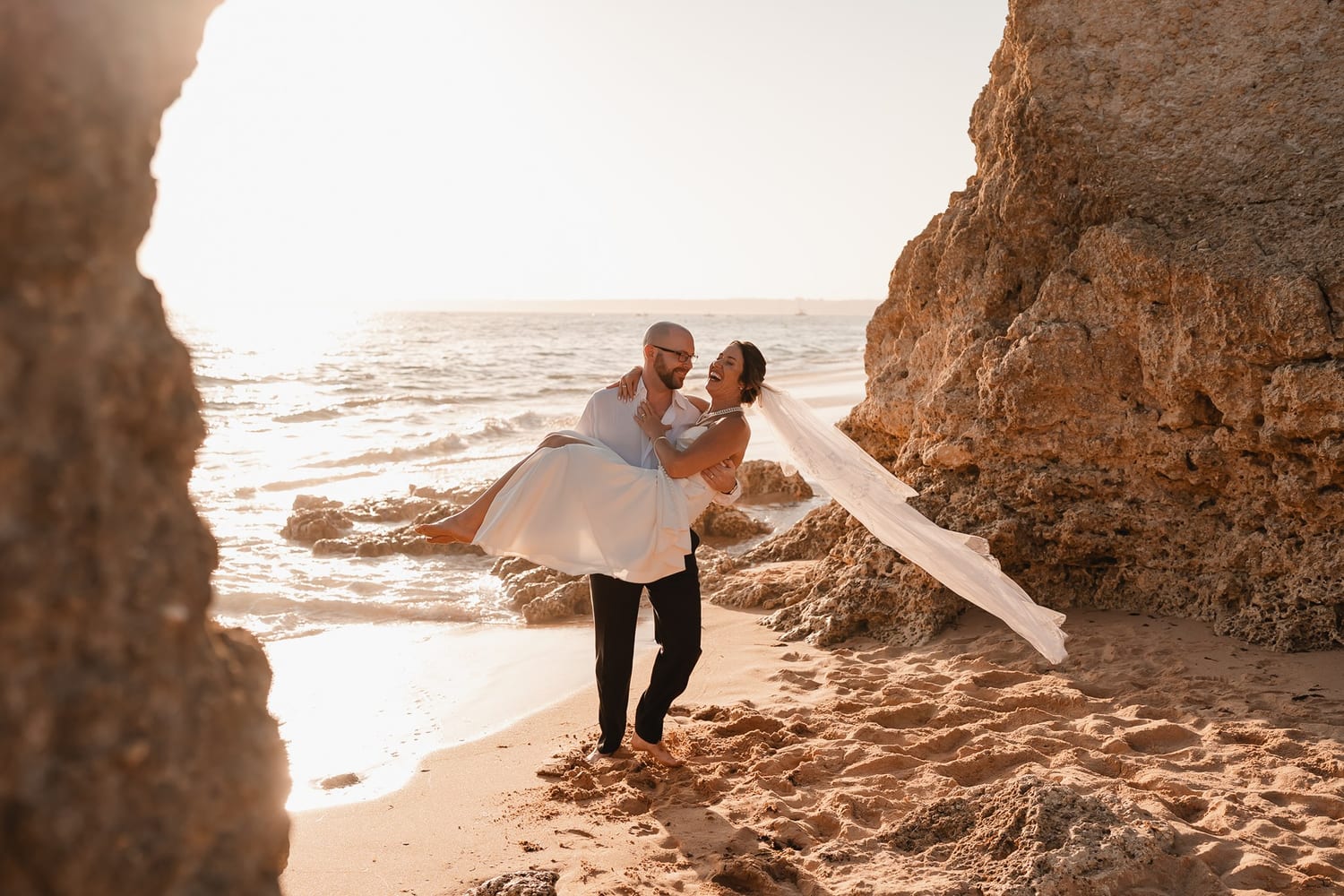 A groom carrying his bride along a sandy cove between golden rock walls