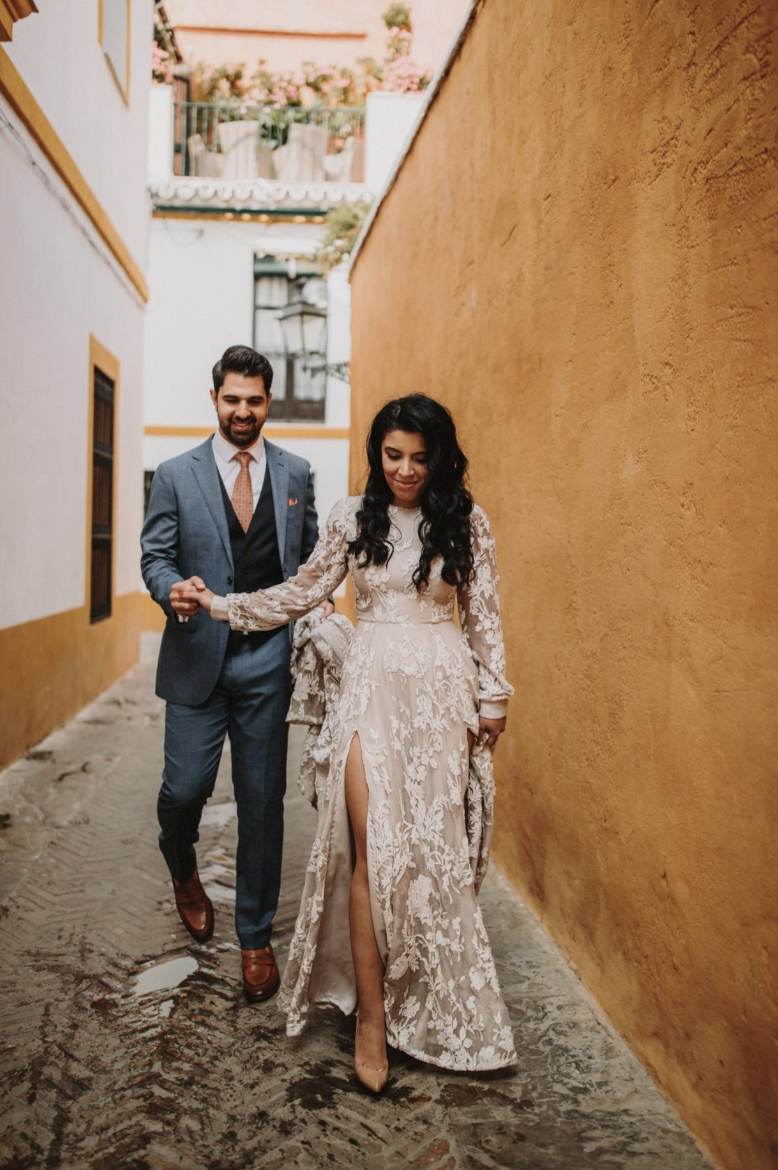 A woman in a lace wedding dress walking with a man in a blue suit in a narrow alleyway with yellow and white walls.