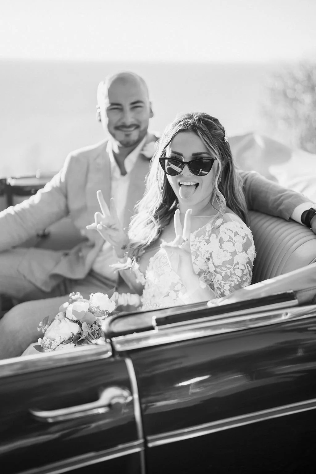 A woman in sunglasses and a lace dress making peace signs while sitting in a car with a man in a light suit smiling in the background.