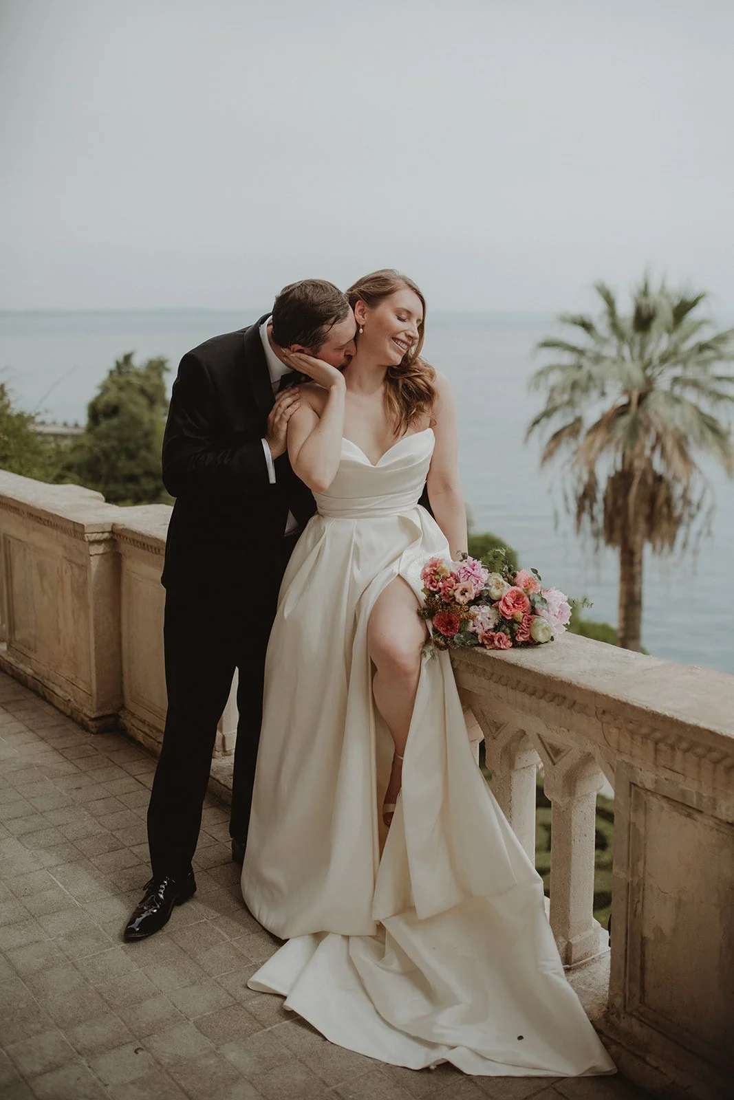 A newlywed couple sharing an intimate moment on a balcony overlooking the ocean, with a palm tree in the background. The bride is seated on the balcony railing, holding a bouquet of pink and white flowers, wearing a white wedding gown. The groom, dressed in a black tuxedo, is leaning in close to her, whispering or kissing her cheek.