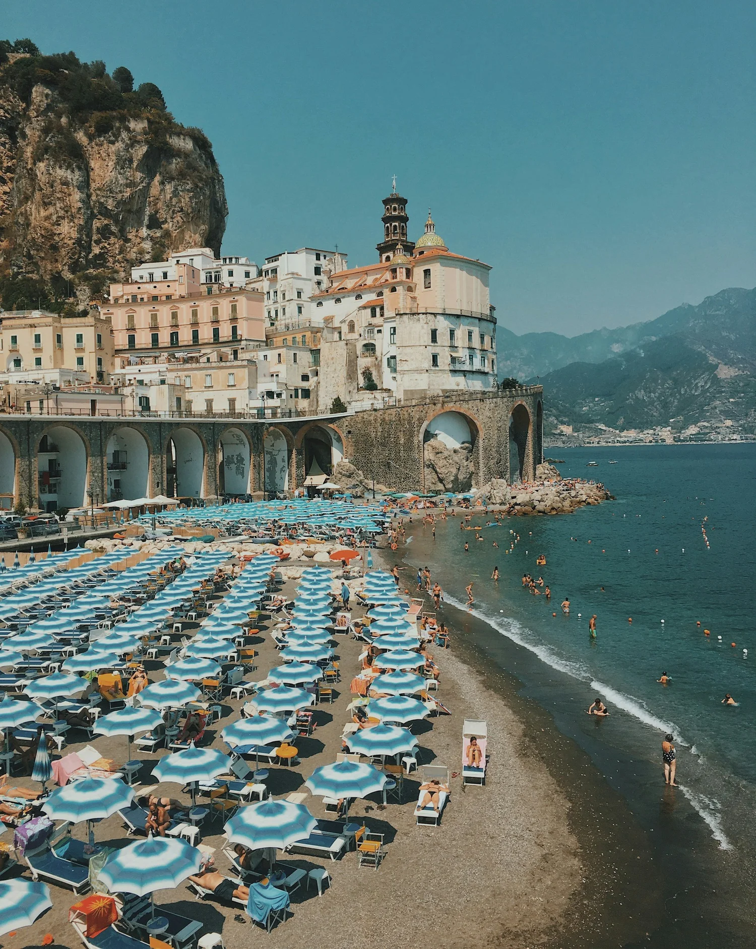 Beach filled with numerous blue and white striped umbrellas, people swimming and relaxing in the water, and a backdrop of buildings, a church, rocky cliffs, and mountains.