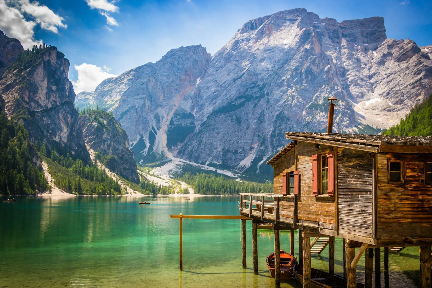 A wooden house with red window shutters by a clear green lake, surrounded by tall mountains with a partly cloudy blue sky.