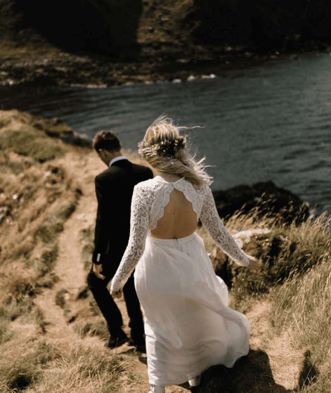 Bride in a white lace wedding gown with open back walking on a grassy path near the ocean, accompanied by a groom in a black suit, during a scenic outdoor wedding near cliffs.