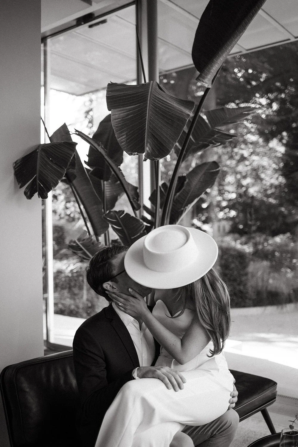 A black and white photo of a couple sharing a kiss indoors, with a large potted plant and window in the background. The woman wears a wide-brimmed hat and a sleeveless dress, while the man wears a suit and glasses.