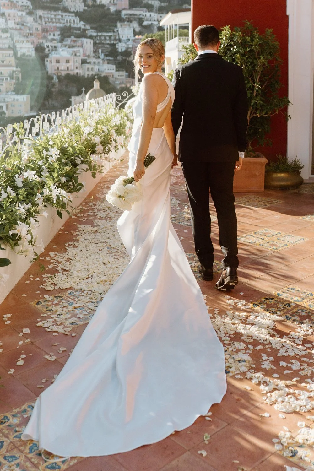 A bride in a white wedding gown holding a bouquet of white flowers, smiling as she walks away from a groom in a black tuxedo on a decorated outdoor terrace, with flower petals on the floor and a scenic hillside town in the background.