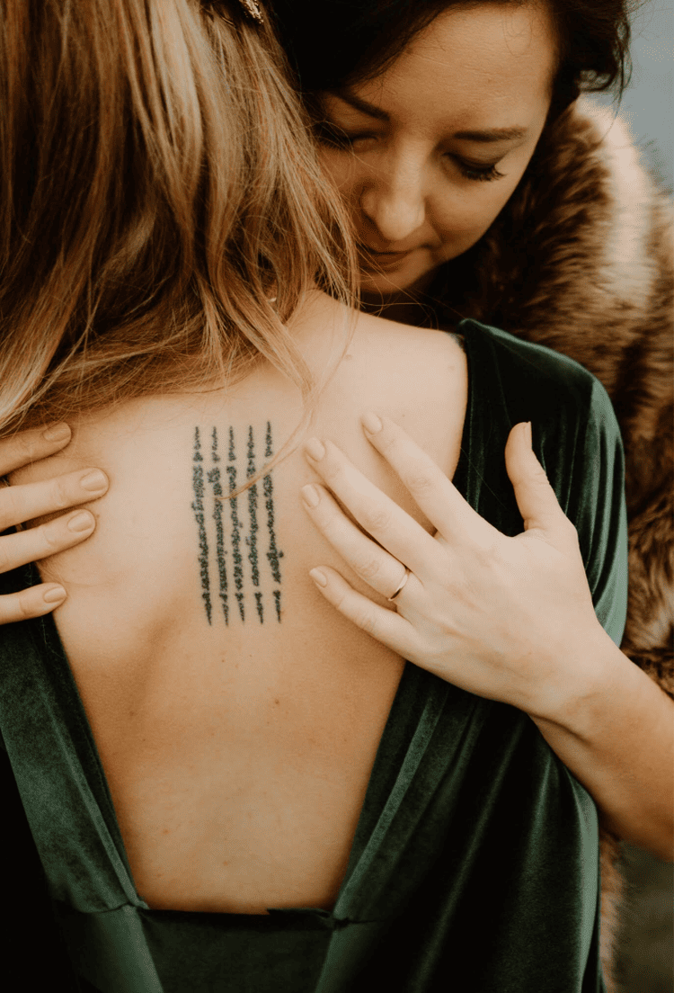 Close-up of two women hugging, one showing a tattoo on her back, with the woman facing away having blonde hair and the woman facing the camera with dark hair and a fur coat.