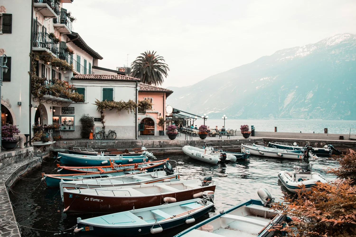 A scenic lakeside scene with boats docked in the water, a row of colorful buildings with balconies and flowers, and mountains in the background.