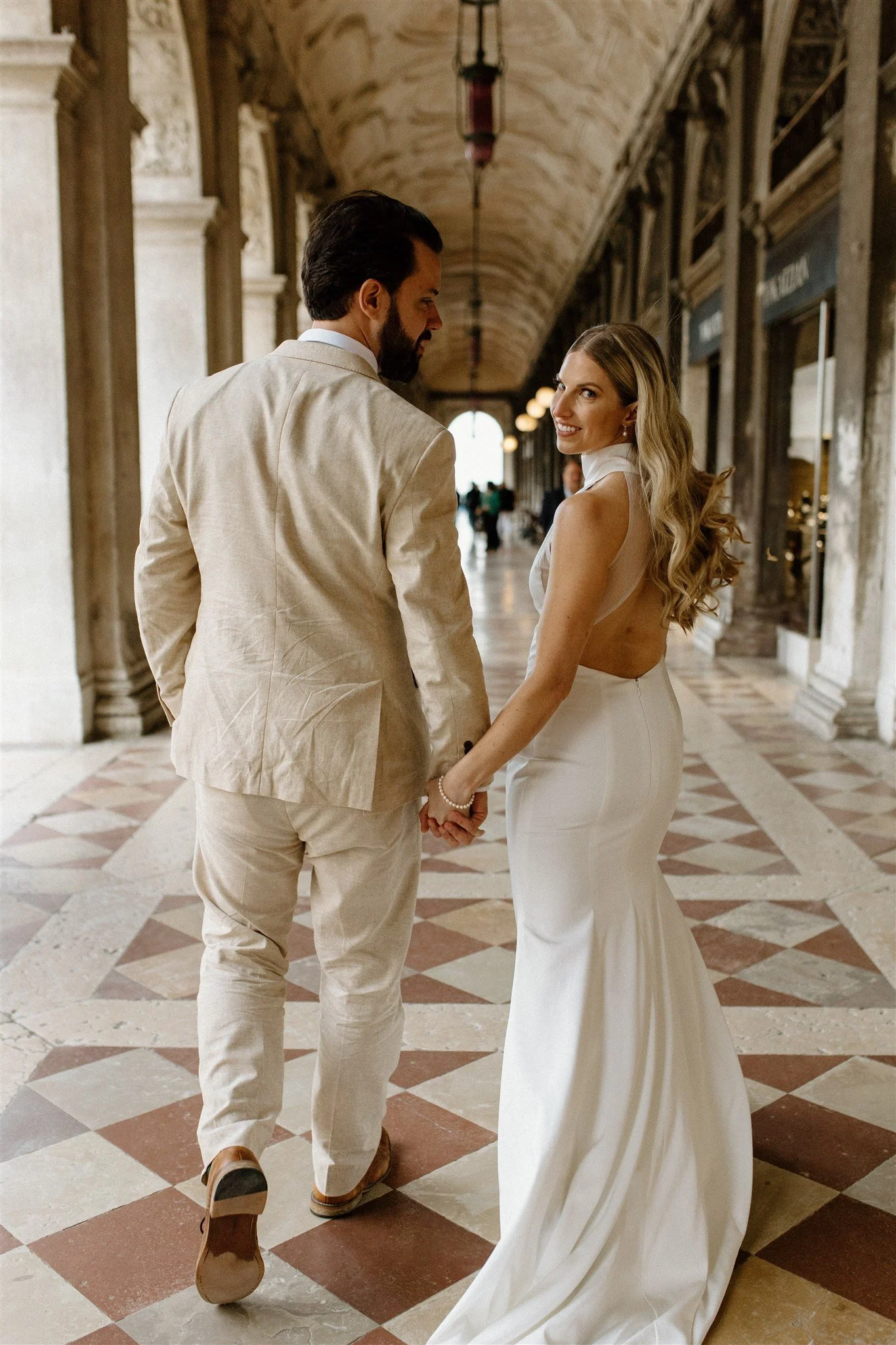 A couple dressed in wedding attire holding hands and looking at each other in a historic, ornate indoor hallway with high arched ceilings and checkered tile floors.