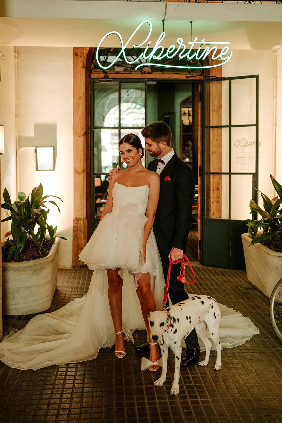 A newlywed couple stands indoors, the bride in a strapless white wedding dress and the groom in a black tuxedo. They are smiling with the groom holding a leash attached to a Dalmatian dog. A neon sign above reads