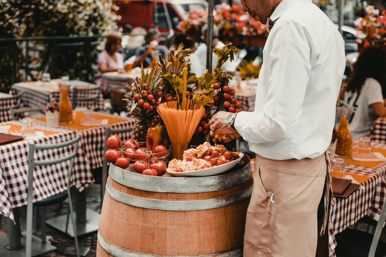 A man in a white shirt and beige apron arranging tomatoes and floral decorations on a barrel at an outdoor market with checkered tablecloths and other people in the background.