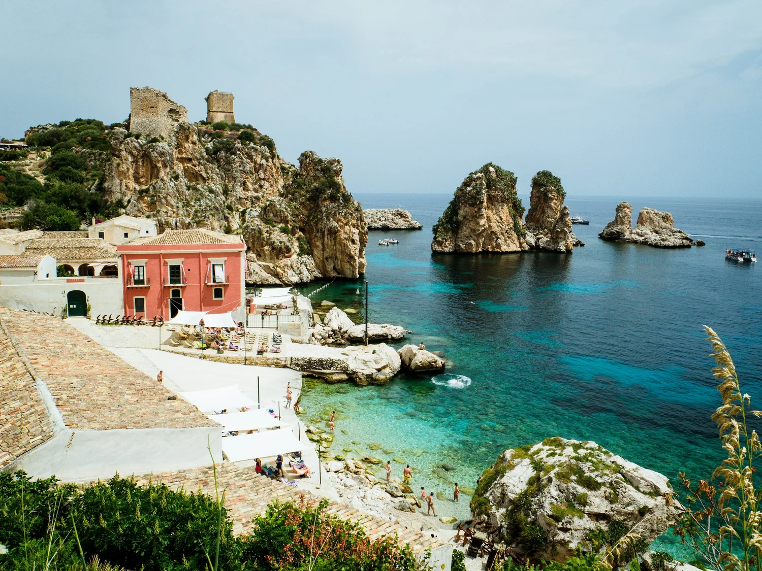 A coastal scene with rocky cliffs, colorful buildings, and clear blue water, featuring a beach area with sunbathers and boats in the bay.