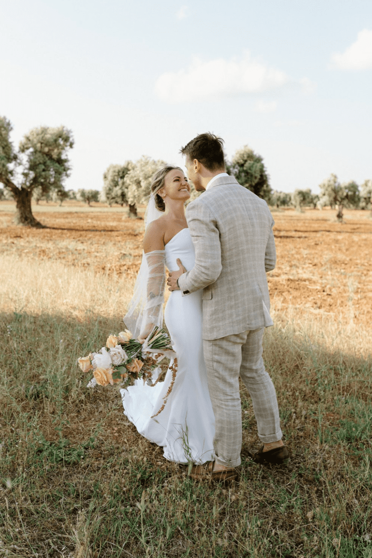 A bride and groom standing in a field of grass and trees, smiling and looking at each other, with the bride holding a bouquet of flowers.