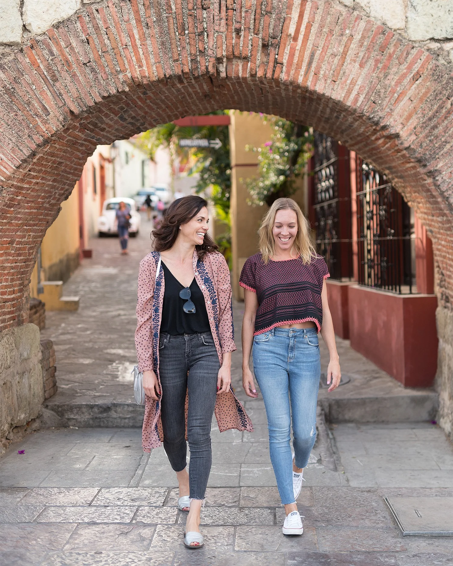 Two women walk together smiling beneath a brick archway on a cobblestone street in a colorful town with buildings, people, and cars in the background.