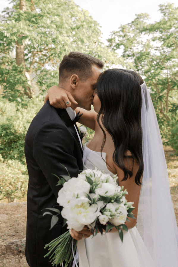 Bride and groom kissing outdoors, bride holding a bouquet of white flowers, bride has long dark hair, groom in a tuxedo with a bow tie, trees in the background.
