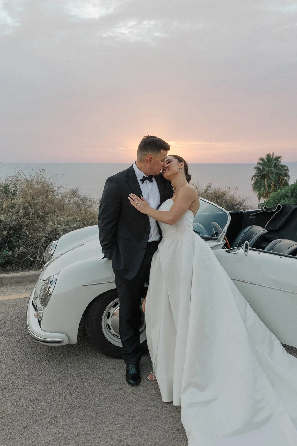 A newlywed couple in wedding attire, a man in a tuxedo and a woman in a white wedding gown, sharing a romantic moment beside a classic white convertible car with the sunset over the ocean in the background.