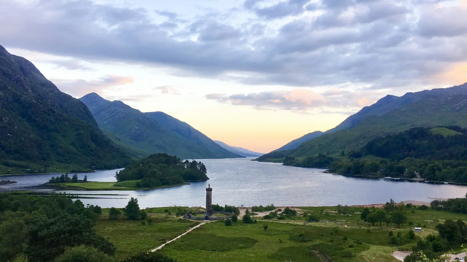 Scenic view of a river flowing through a valley surrounded by green hills and mountains, with some clouds in the sky at sunset.