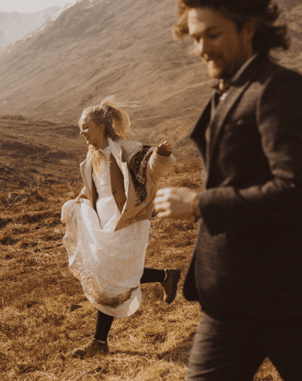 A woman and a man running outdoors in a mountainous field during sunset, with the woman wearing a white dress and the man in a dark suit.