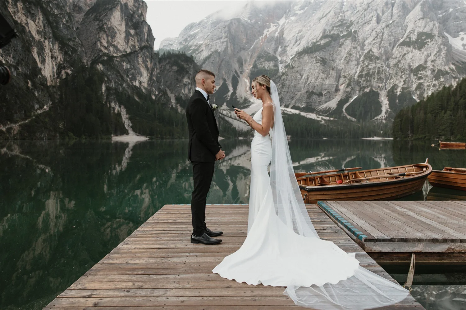 A bride and groom standing on a wooden dock by a lake surrounded by mountains, with two wooden boats nearby. The bride is wearing a white wedding dress and a veil, and the groom is in a black tuxedo. The bride is looking at her phone while the groom faces her, with a mountain landscape in the background.